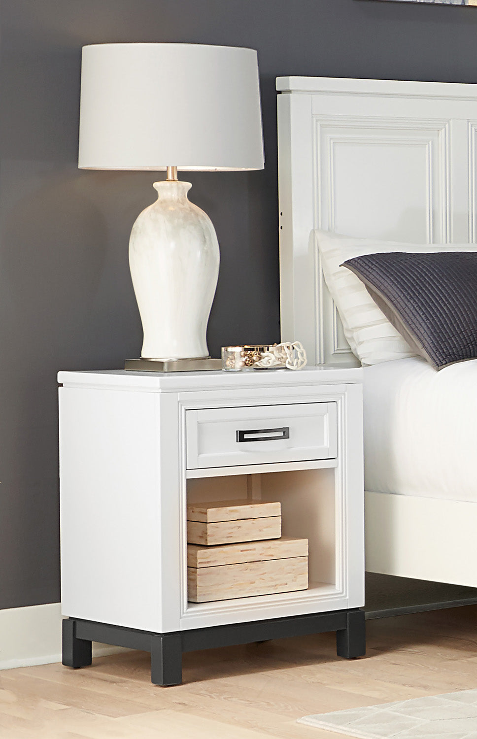 White nightstand with a lamp and decorative items next to a bed in a bedroom setting.