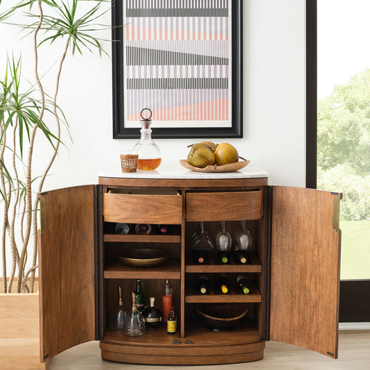 Wooden bar cabinet with open doors displaying bottles and glasses, with a plant and framed picture in the background.