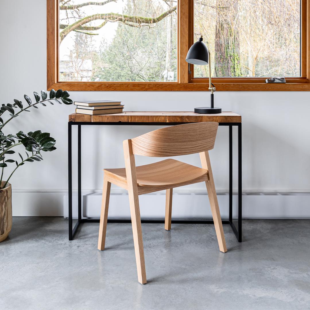 Wooden chair in front of a desk with a lamp and books, in a room with large windows.