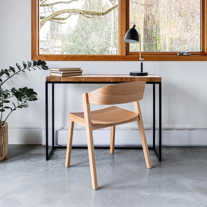 Wooden chair in front of a desk with a lamp and books, in a room with large windows.
