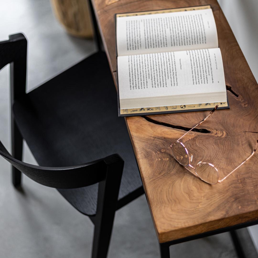 Open book on a wooden table with glasses next to it, black chair in the foreground
