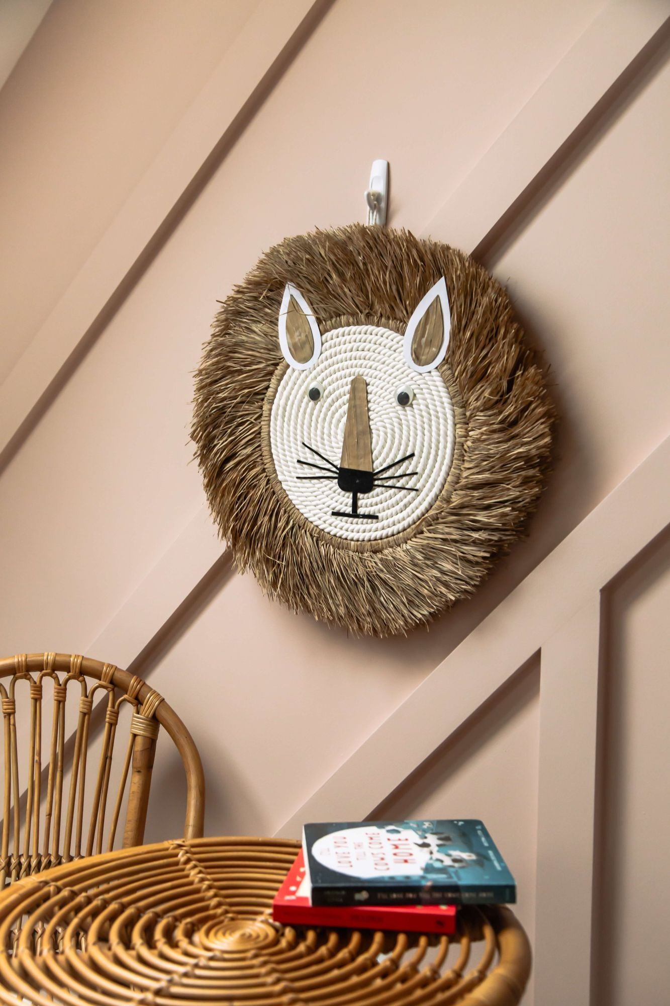 Woven lion head wall decoration on a beige wall with a wicker chair and books below.