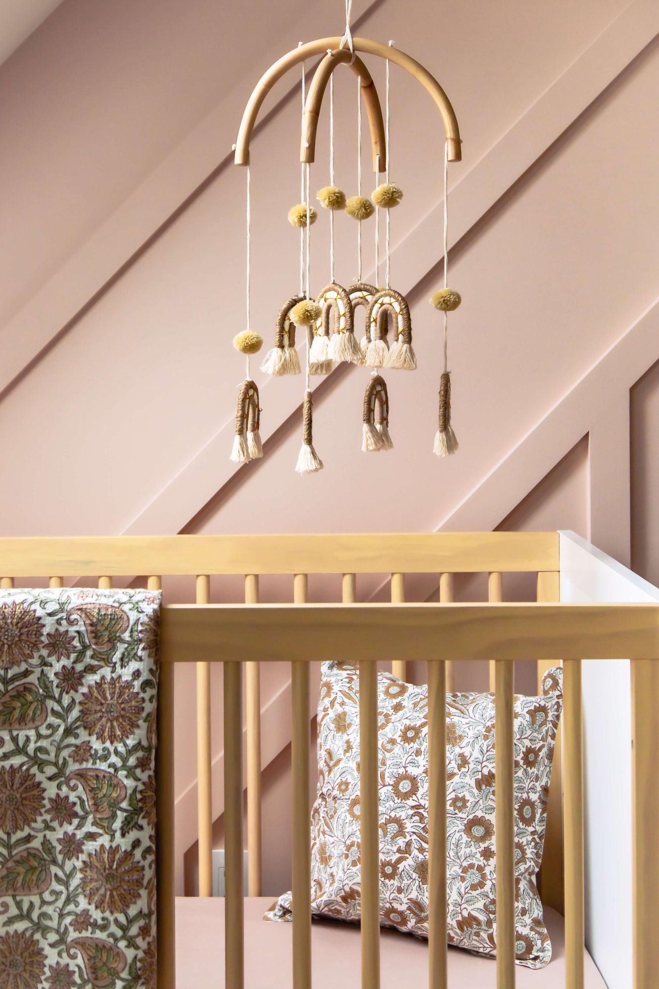 Baby crib with decorative pillows and a hanging mobile against a pink wall.