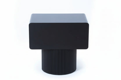Black cube-shaped stool with ribbed cylindrical base on a light wooden floor.