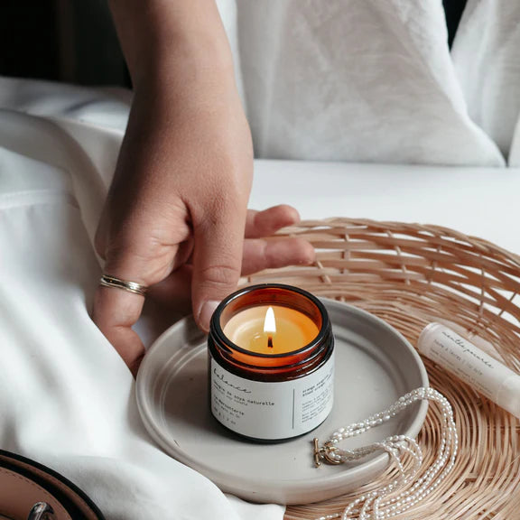 Hand lighting a candle in a small jar on a white surface with a wicker basket in the background.