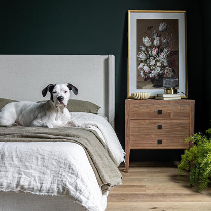 Dog lying on a bed in a bedroom with a wooden nightstand and floral artwork.