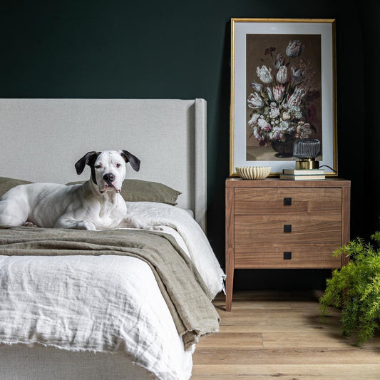 Dog lying on a bed in a bedroom with a wooden nightstand and floral artwork.