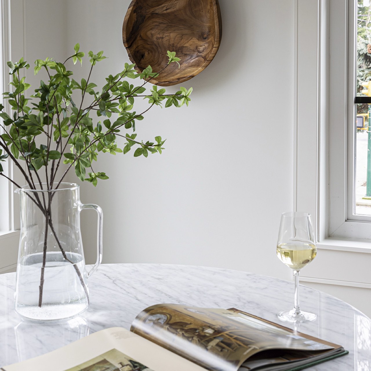 Clear vase with green branches, open book, and glass of white wine on a marble surface.