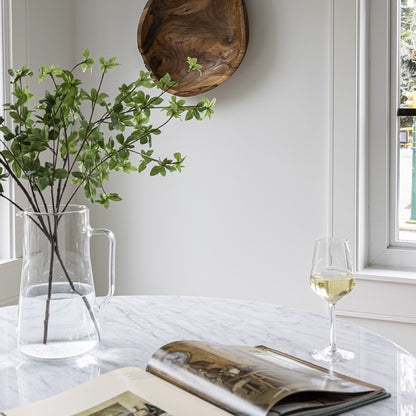 Clear vase with green branches, open book, and glass of white wine on a marble surface.