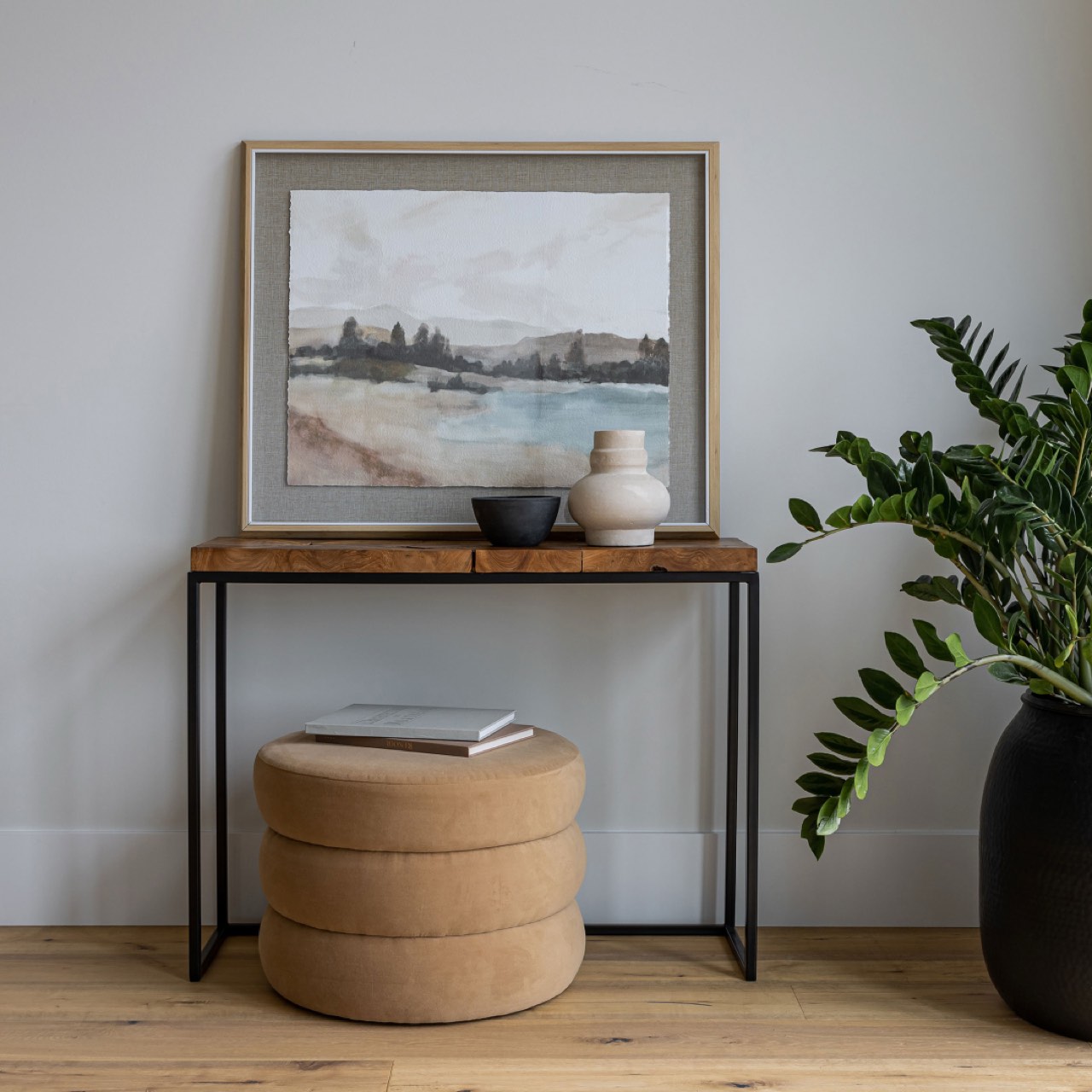 Wooden side table with a framed landscape painting, books, and a vase in a room with a plant.