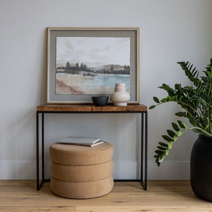 Wooden side table with a framed landscape painting, books, and a vase in a room with a plant.