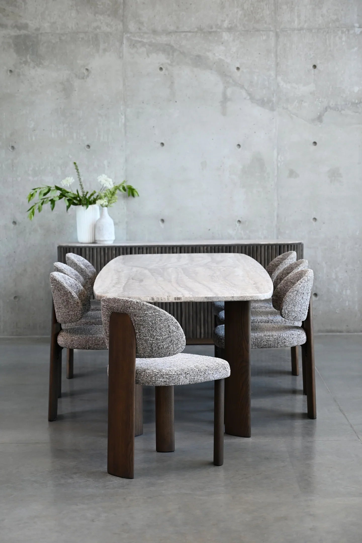 Dining table with marble top and wooden legs, surrounded by chairs with patterned cushions, against a concrete wall.