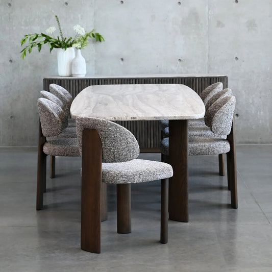 Dining table with marble top and wooden legs, surrounded by chairs with patterned cushions, against a concrete wall.