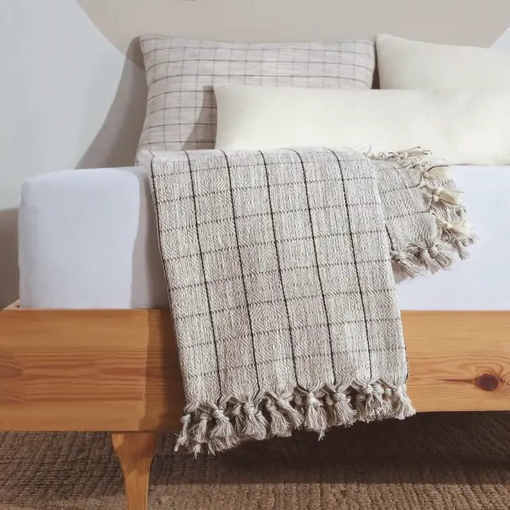 Textured blanket with tassels draped over a wooden bench against a neutral wall.